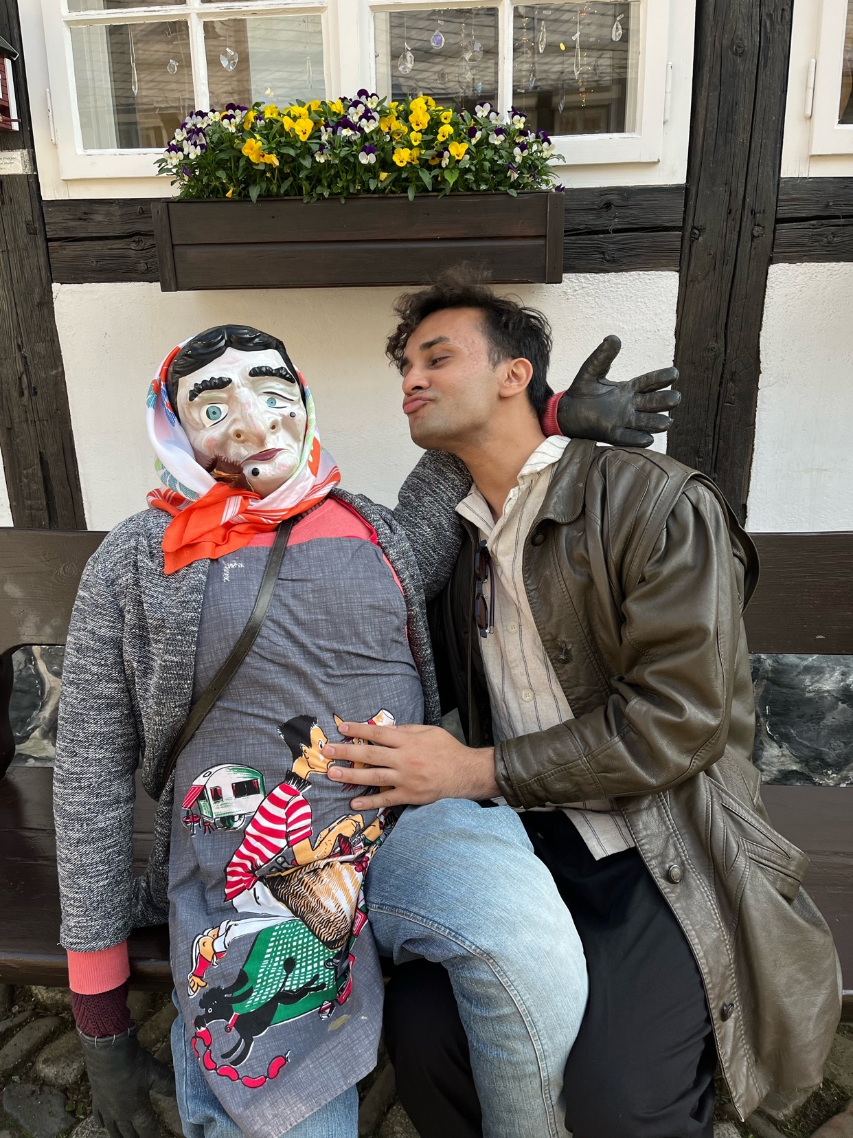 A man poses with a doll, leaning in for a kiss. He wears a leather jacket and a stylish outfit.