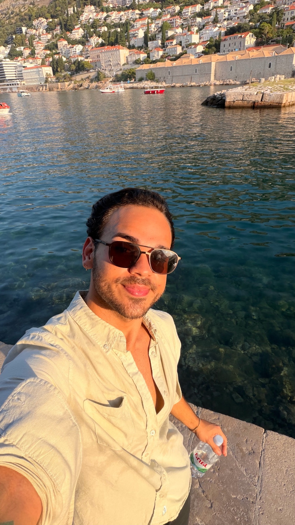 A man takes a selfie by the water with a coastal city in the background. He is wearing sunglasses and a light-colored shirt.