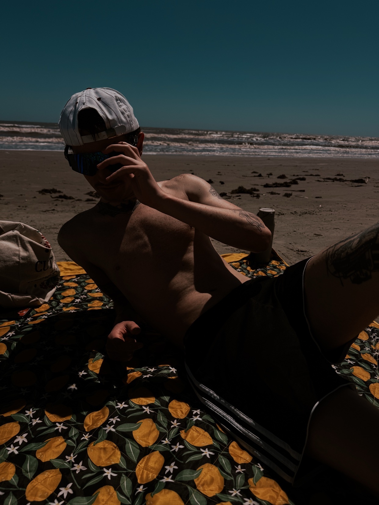 A man wearing a white baseball cap and sunglasses lies on a beach towel on the sand. He is looking towards the camera, holding a phone or camera up to his face. The ocean and sky are visible in the background.