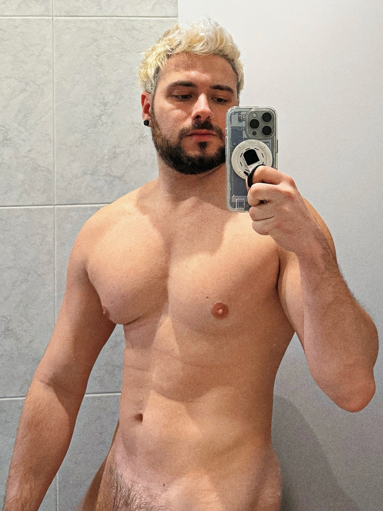 A shirtless man takes a mirror selfie, showcasing his physique. He has blonde hair and a beard.