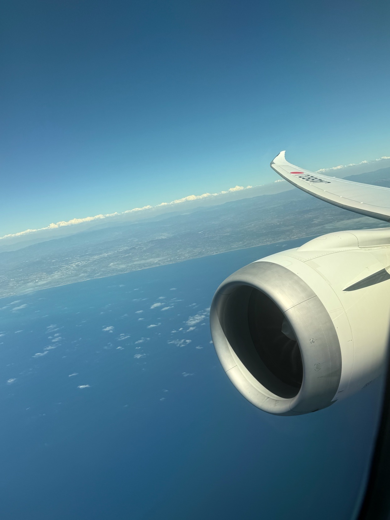 View from an airplane window shows the deep blue ocean below, with a coastline and clouds visible in the distance. The airplane's engine and wing are prominent in the foreground.