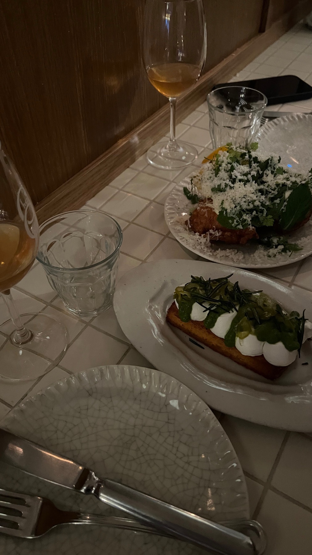 A close-up shot of two plates of food and two glasses of wine on a tiled table. The food appears to be gourmet appetizers, artfully plated with fresh ingredients.