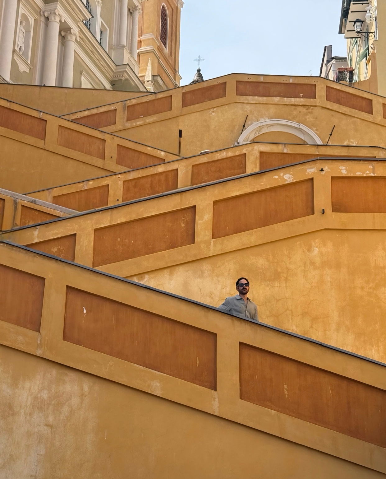 A man stands on a wide, yellow staircase with ornate railings. The architecture suggests a European city.