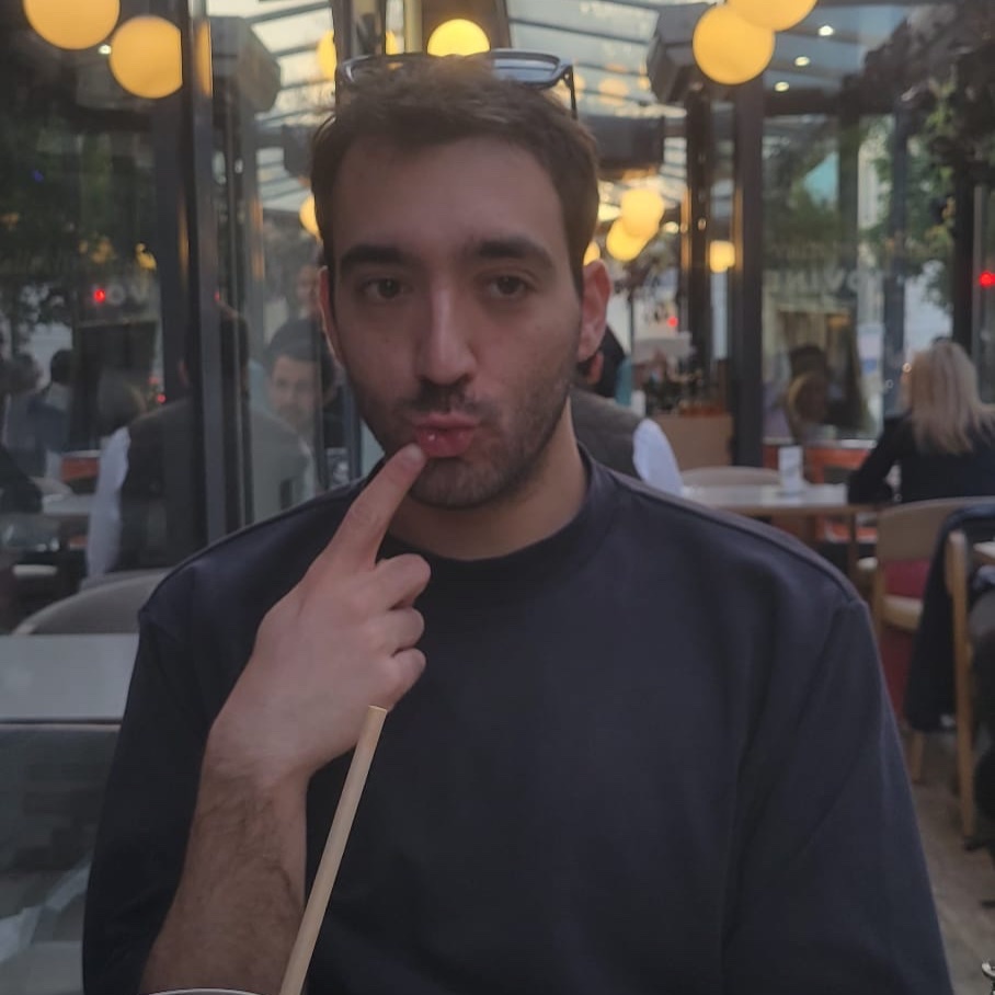 A man with dark hair and stubble pouts with his finger to his lips. He is wearing a dark crew neck shirt and is seated in what appears to be a restaurant.