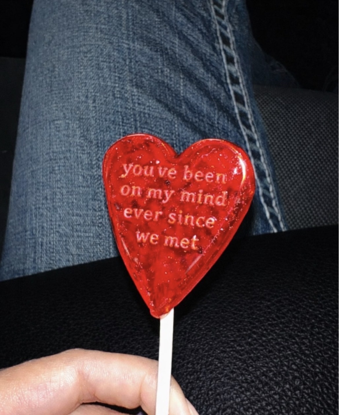 A close-up shot of a red heart-shaped lollipop with the words "You've been on my mind ever since we met" engraved on it. The lollipop is held by a hand, with jeans in the background.