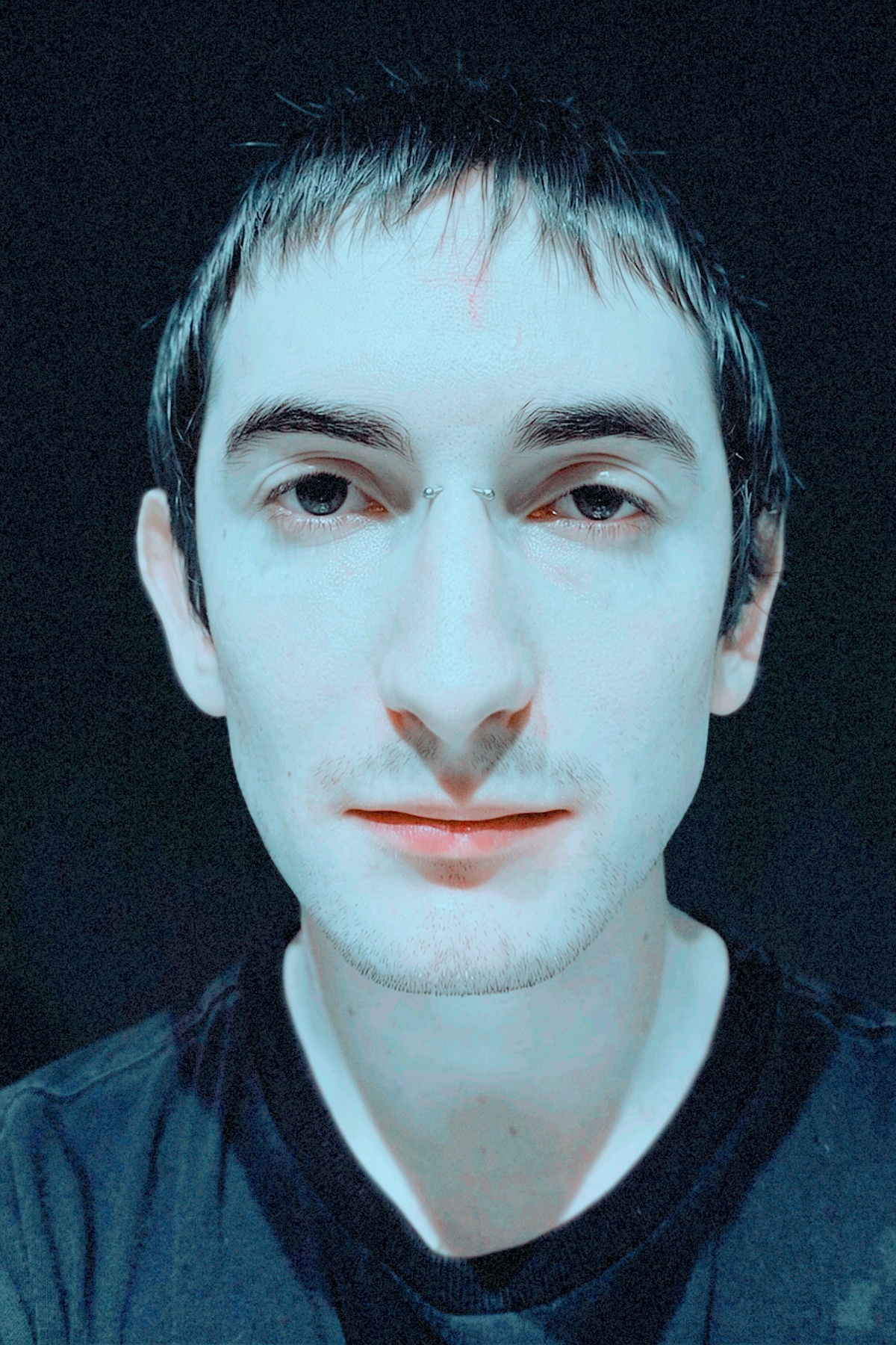 A close-up selfie of a man with dark hair and piercings in his nose and eyebrow. He is wearing a dark t-shirt against a dark background.