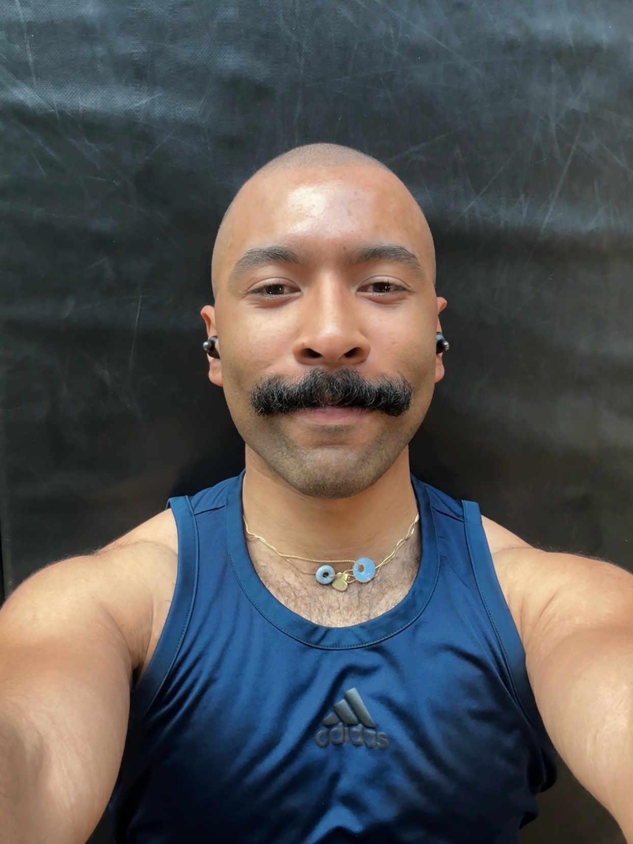 A man with a bald head and a prominent mustache takes a selfie. He is wearing a dark blue tank top with the Adidas logo and a necklace with blue charms.