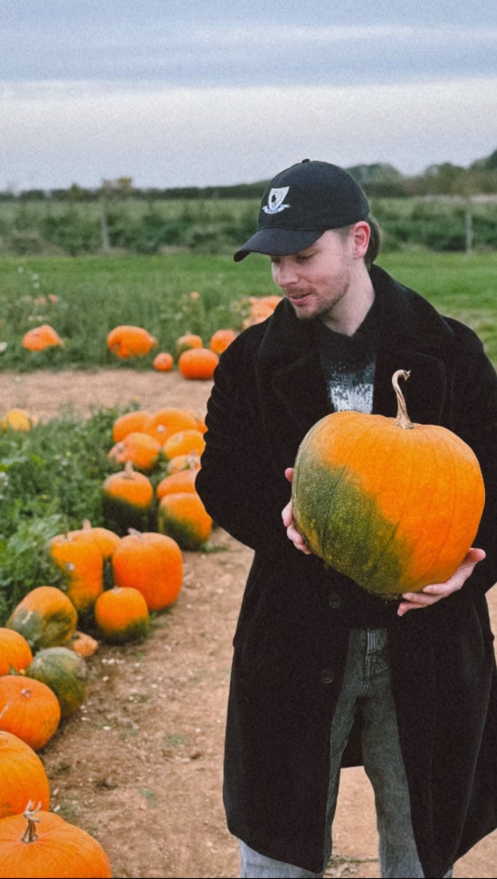 A man holds a large pumpkin in a field. The photo is taken outdoors on a cloudy day.