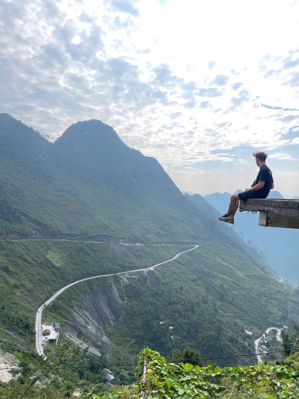 A young man sits on the edge of a concrete platform overlooking a vast, green mountain range with a winding road below. The sky is partly cloudy, and the overall scene evokes a sense of adventure and scenic beauty.