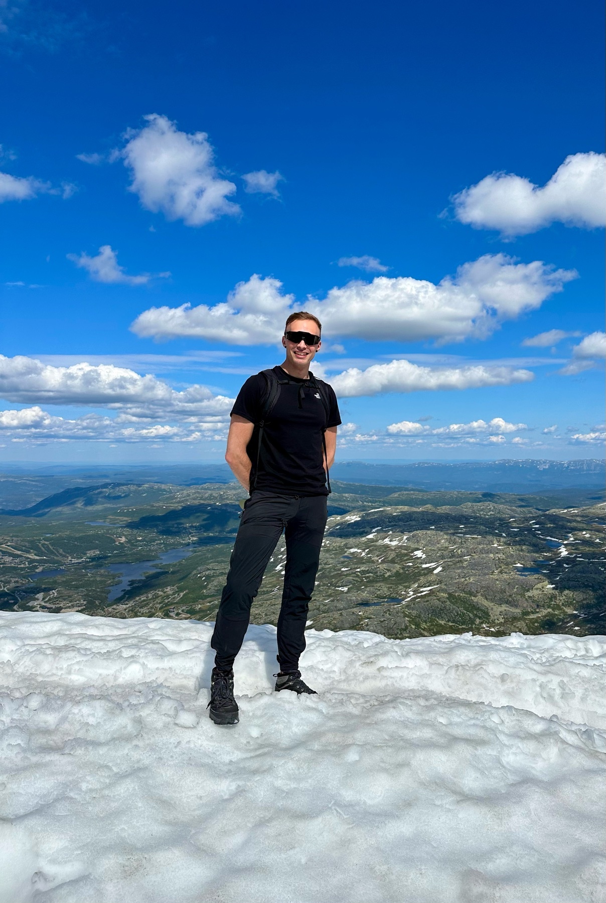 A man stands on a snowy mountain peak, wearing sunglasses and a black t-shirt. The background features a vast landscape under a bright blue sky with fluffy white clouds.