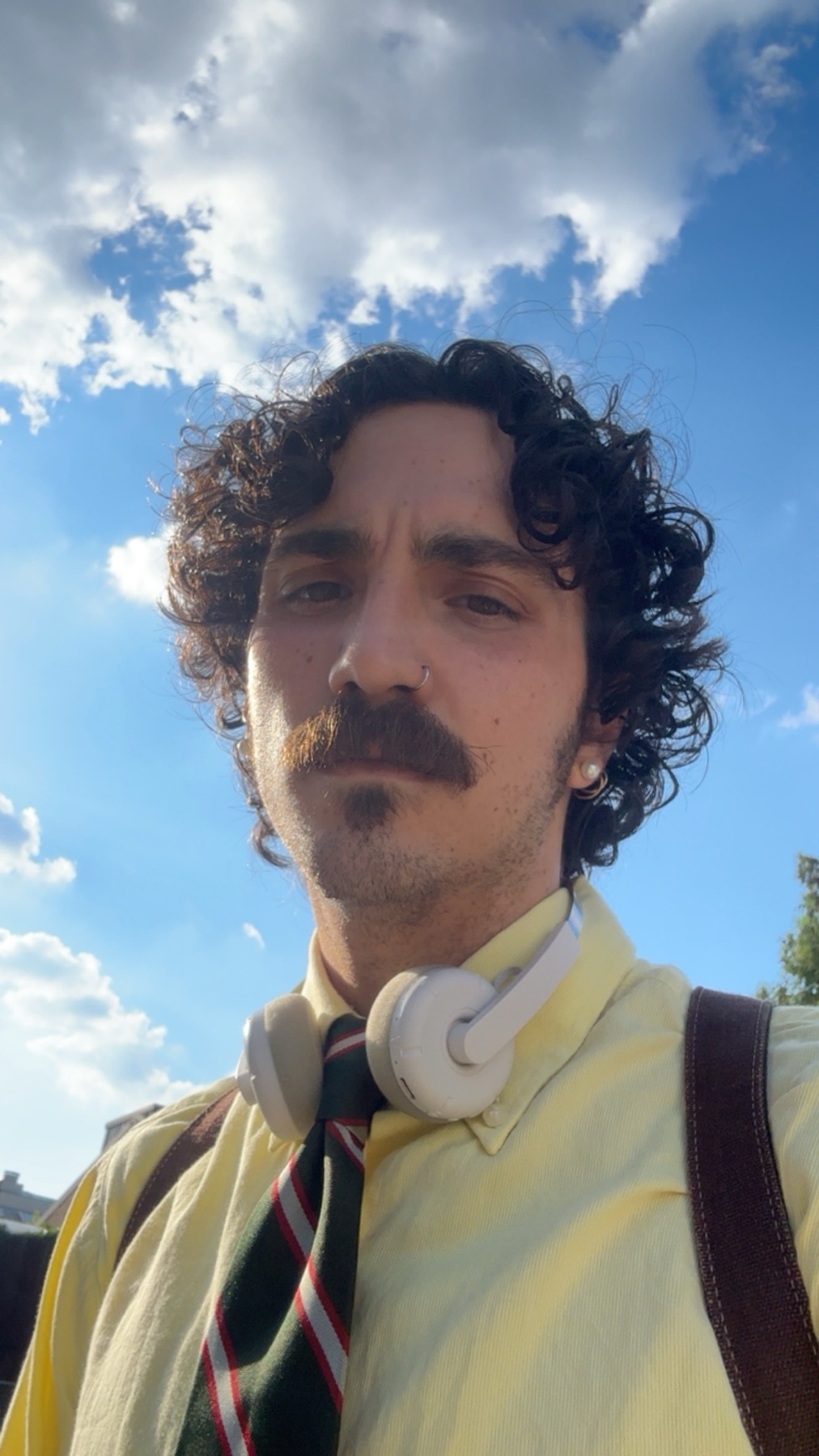 A man with curly dark hair and a mustache poses for a selfie outdoors under a bright blue sky with fluffy white clouds. He is wearing a yellow collared shirt, a striped tie, and headphones around his neck.