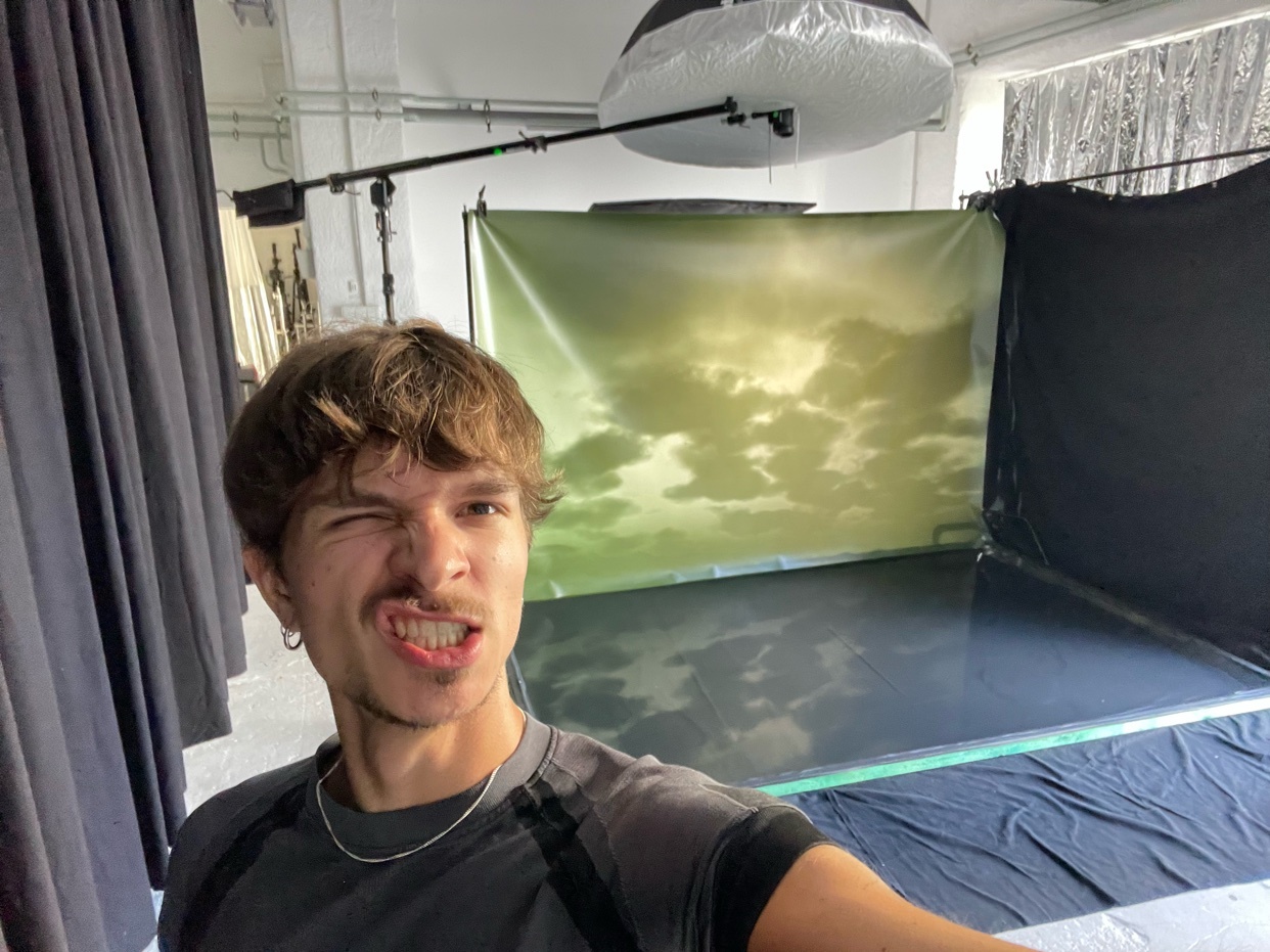 A man takes a selfie in a studio. He is making a funny face and winking at the camera.