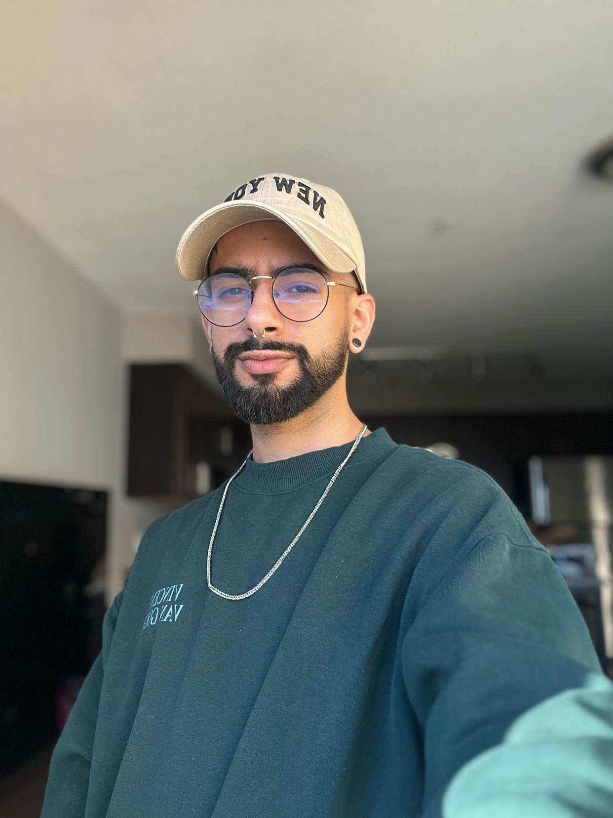 A man wearing a cap and glasses poses for a portrait. He is wearing a green sweatshirt and a silver chain.