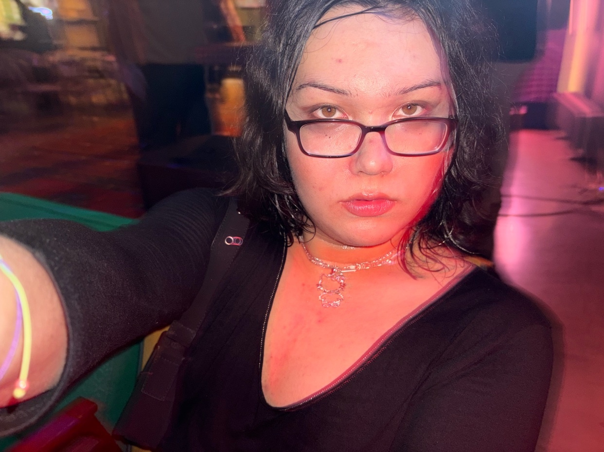A woman takes a selfie indoors. She is wearing glasses and a black top.