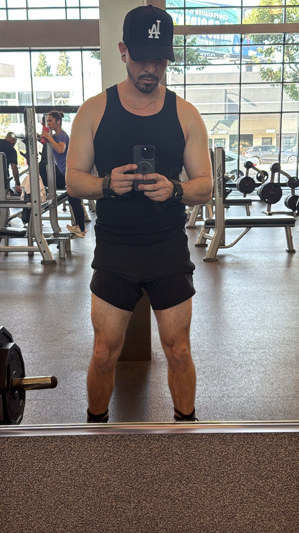 A man takes a mirror selfie at the gym. He is wearing a black tank top, shorts, and a cap.