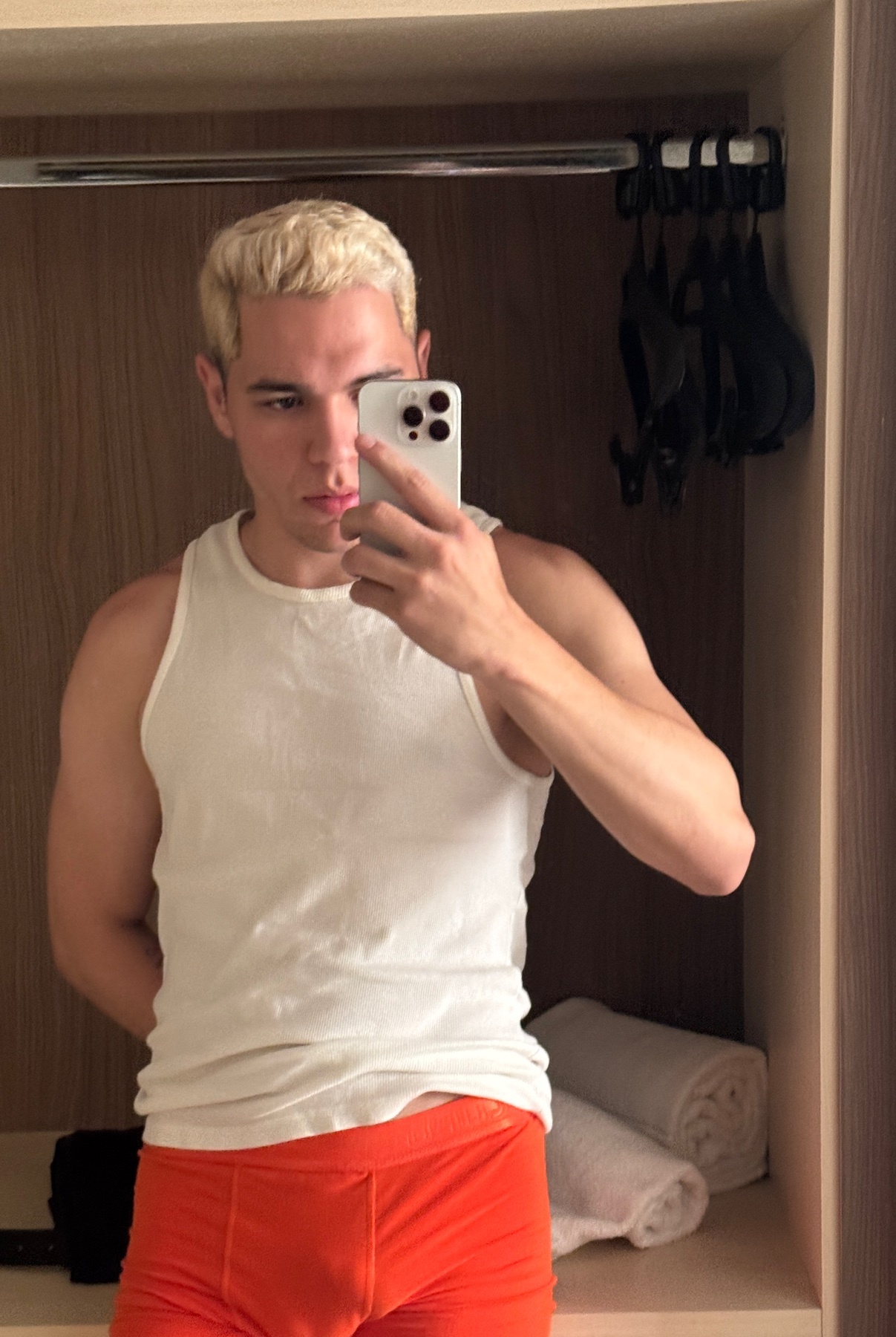 A man takes a mirror selfie, shirtless and wearing orange shorts. He has blonde hair and a muscular physique.