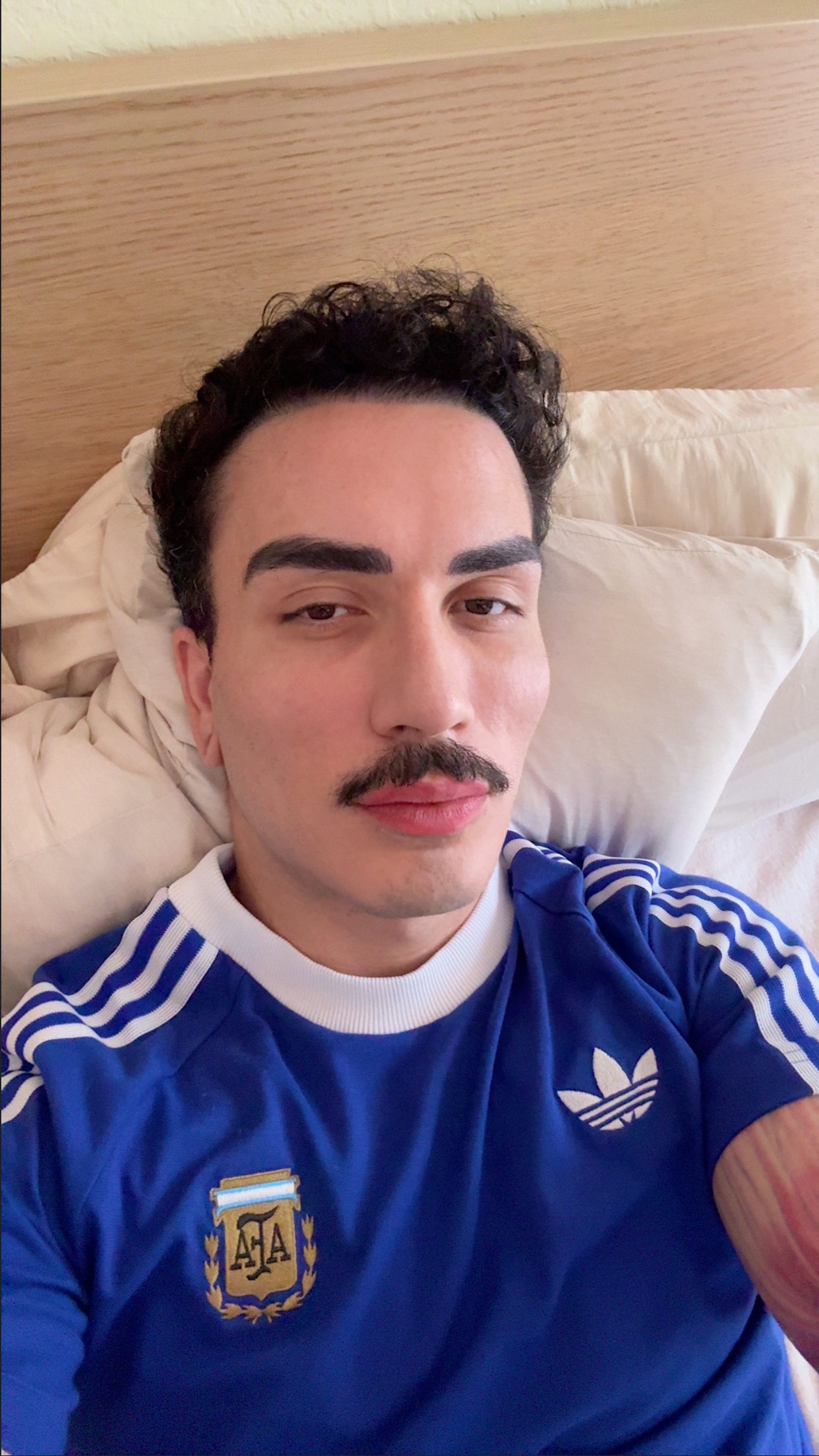 A man with a moustache and curly dark hair lies in bed wearing a blue Adidas jersey with the Argentina crest. He appears to be sick.