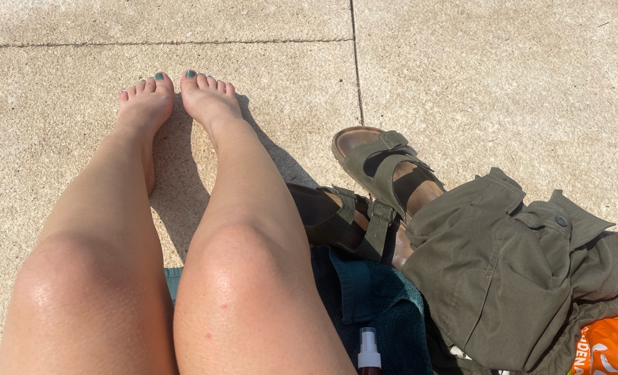 A close-up shot of two people's legs and feet as they relax outdoors. One person's legs are bare and tanned, while the other wears green shorts and Birkenstock sandals.