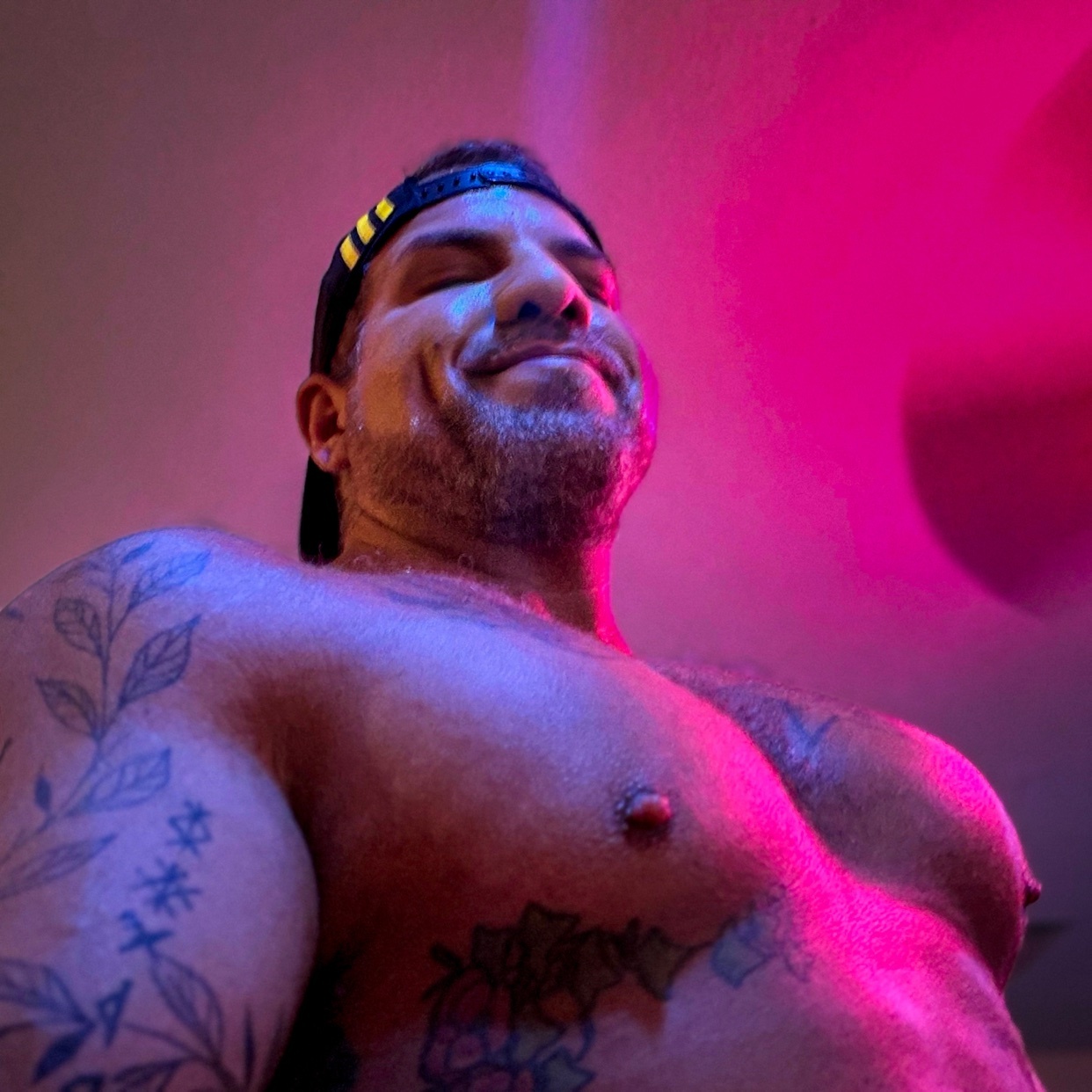 A low-angle shot of a shirtless man wearing a baseball cap, with tattoos on his arms and chest. He has a beard and is smiling.