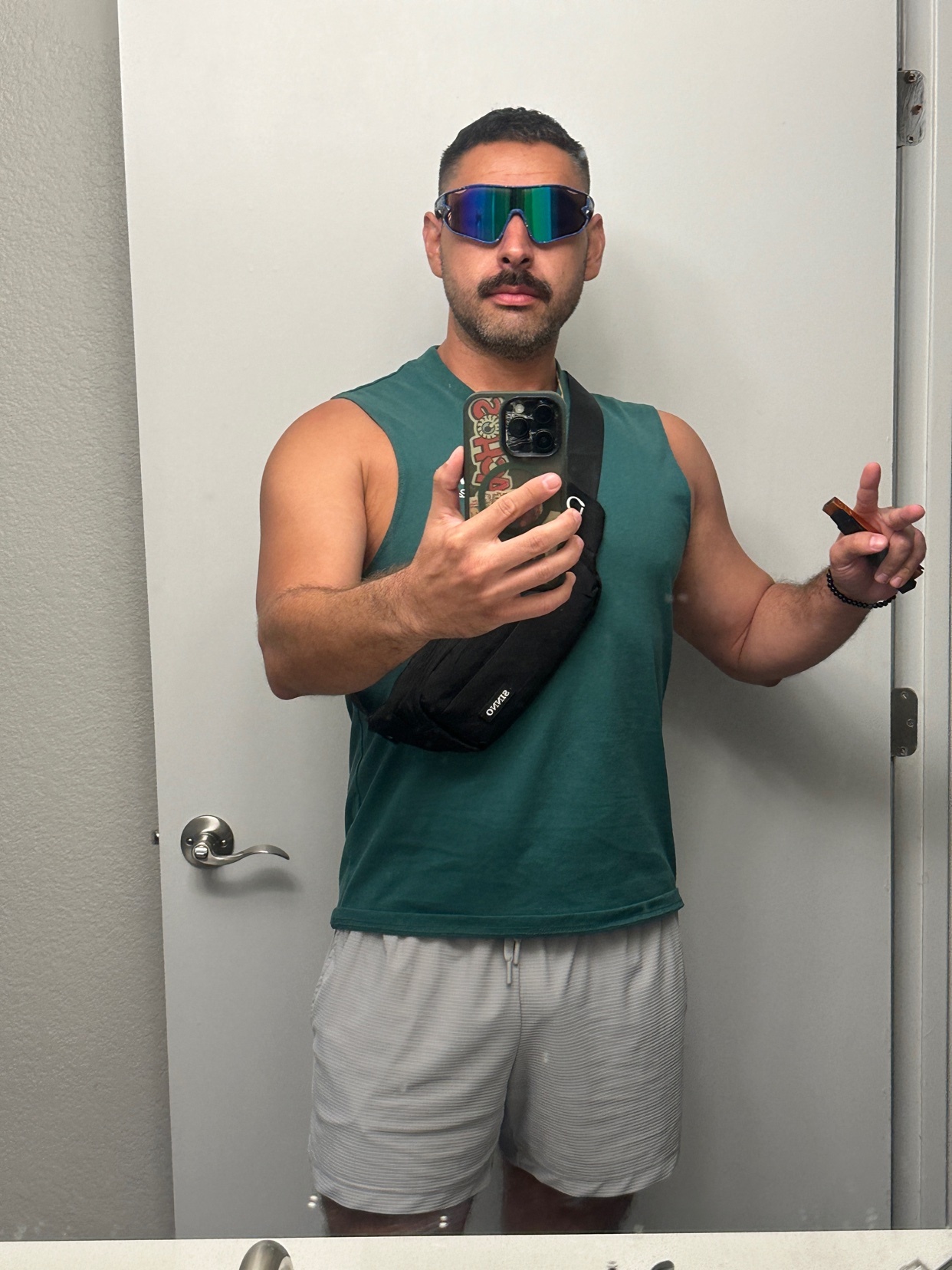 A man with a mustache and sunglasses takes a selfie in a sleeveless shirt and shorts, wearing an arm bag.