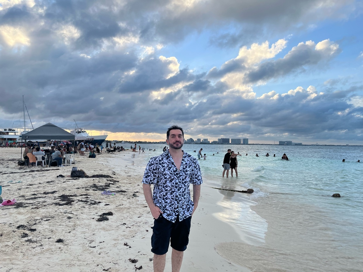 A man stands on a sandy beach with the ocean and a cloudy sky behind him. In the background, there are people swimming, boats, and buildings.
