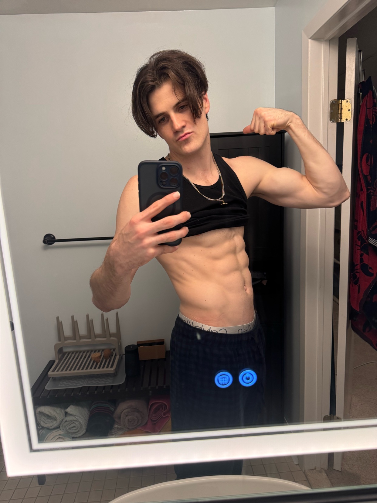 A man flexes his muscles in a mirror selfie. He is shirtless, showing off his muscular physique.