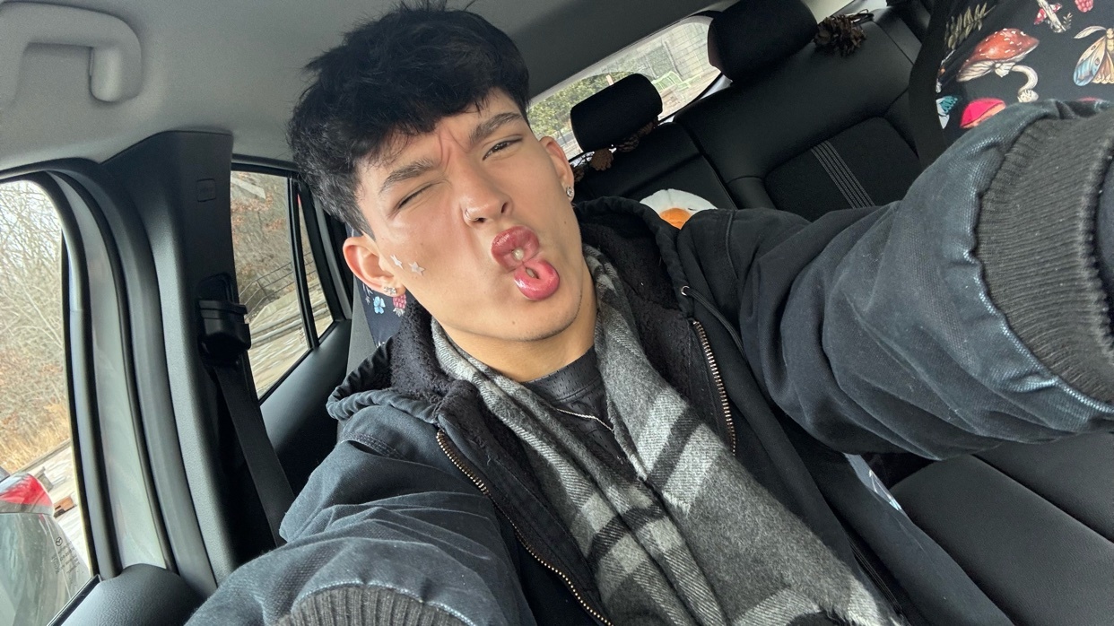 A young man takes a selfie in a car, making a funny face. He is wearing a scarf and a black jacket.