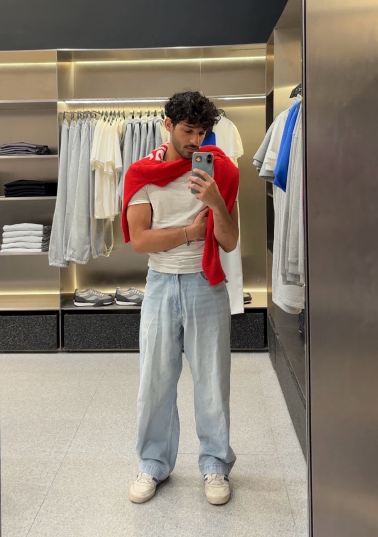 A man takes a mirror selfie in a clothing store, wearing a white t-shirt, light wash baggy jeans, and a red sweater draped over his shoulders. He has dark, curly hair and is wearing sneakers.