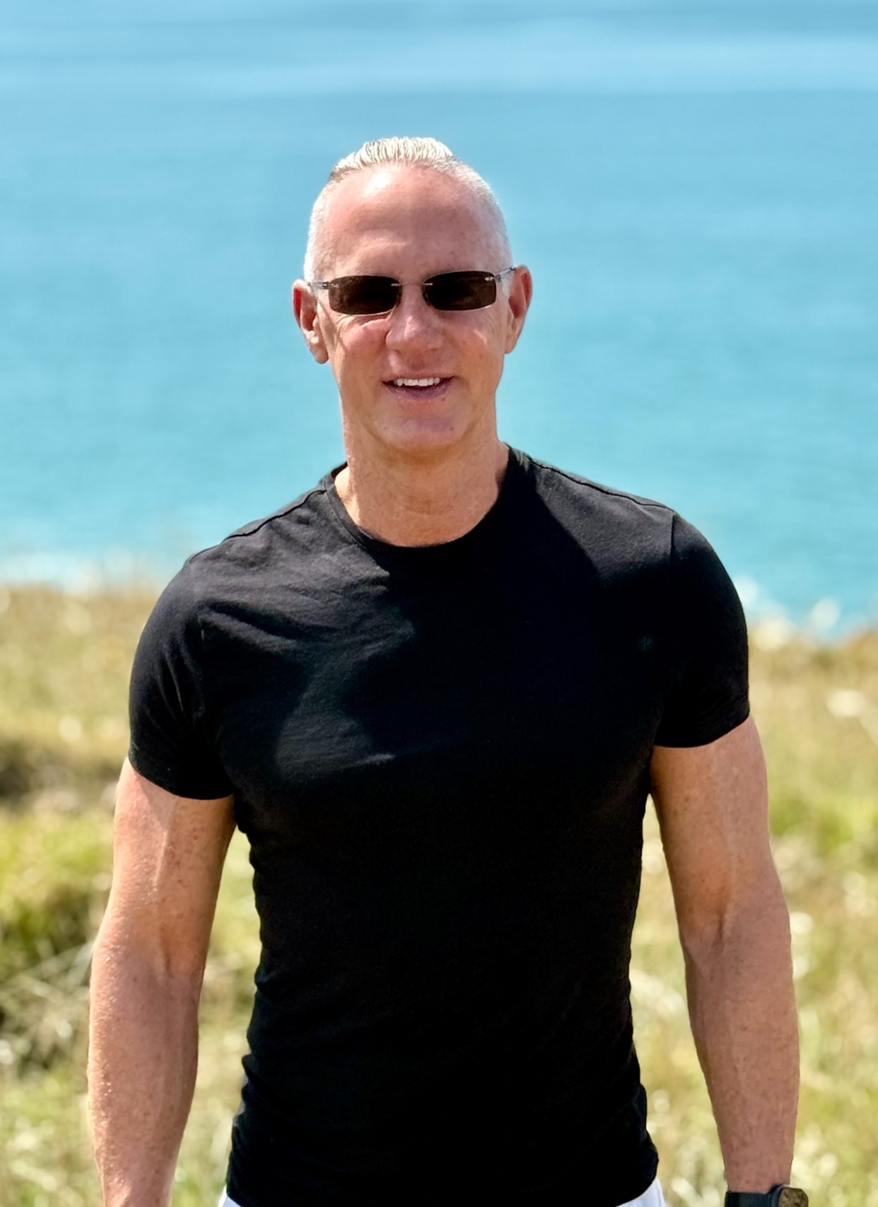 A man with short grey hair and sunglasses poses outdoors wearing a black t-shirt. He appears muscular and is smiling.