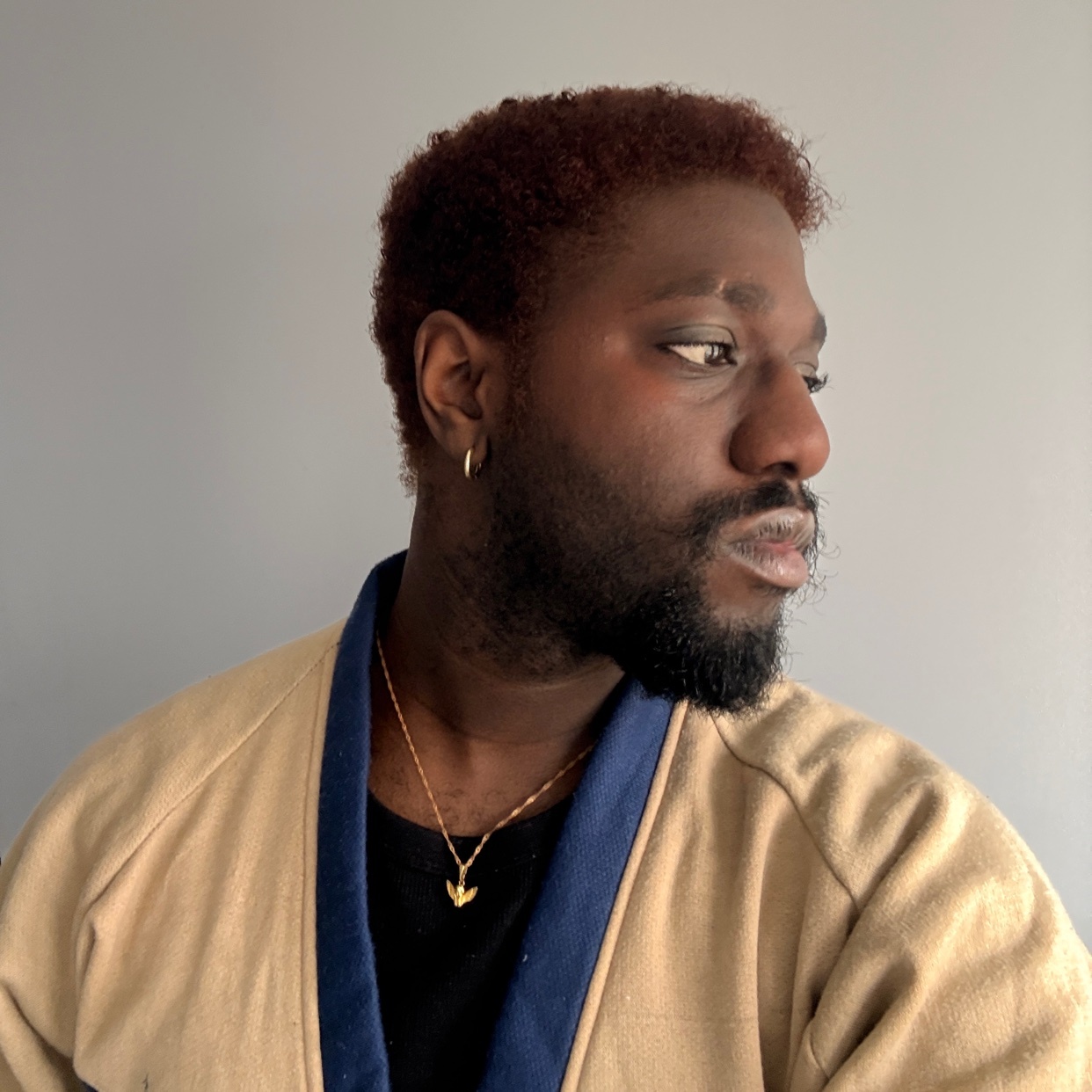 A man with a beard and reddish-brown hair looks off to the side. He is wearing a tan jacket with blue trim over a black shirt and a gold necklace.