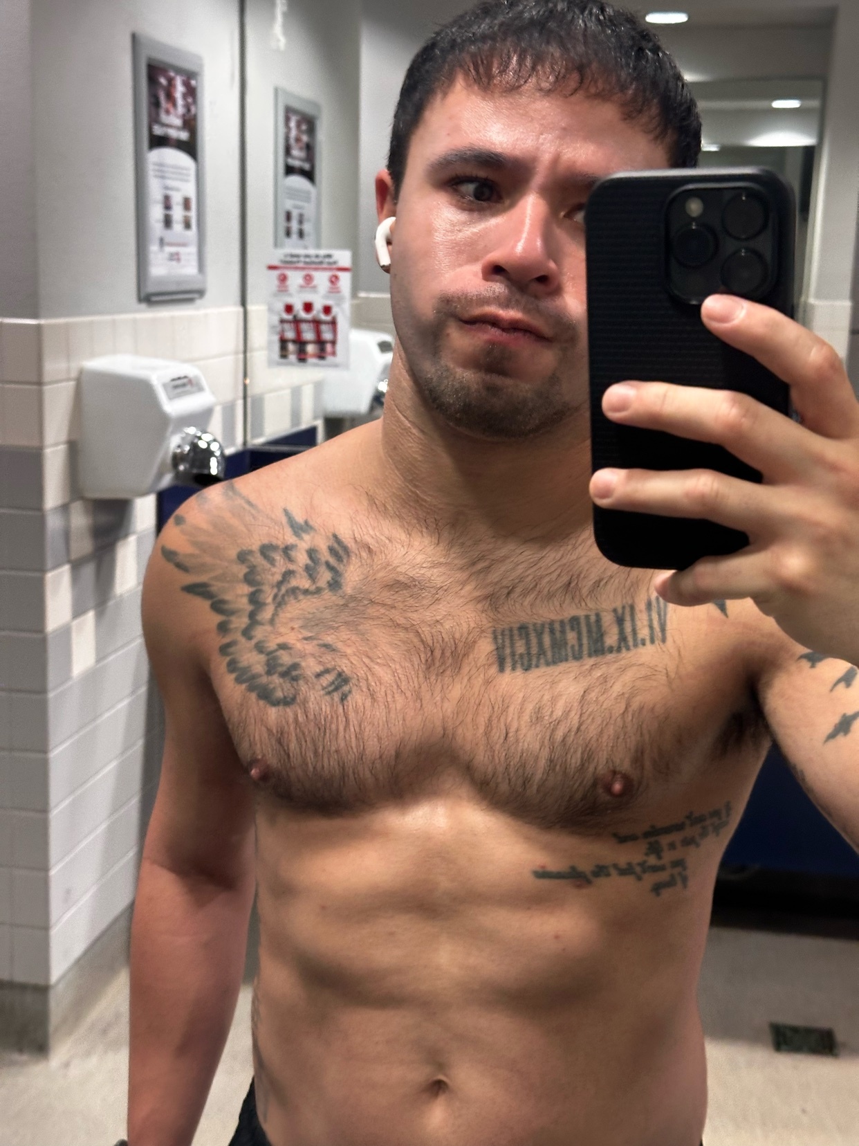 A shirtless man takes a selfie in a gym bathroom. He has tattoos and is wearing earbuds.