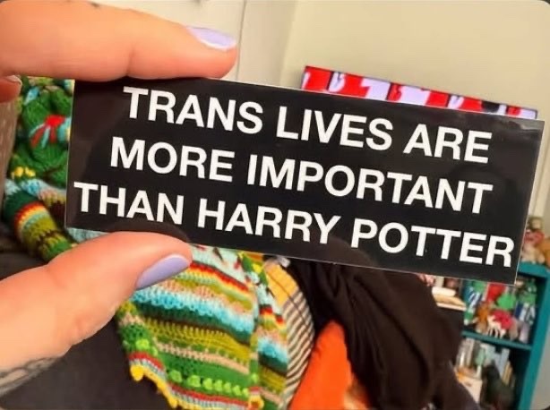 A hand holds up a black sticker with white text that reads "TRANS LIVES ARE MORE IMPORTANT THAN HARRY POTTER".