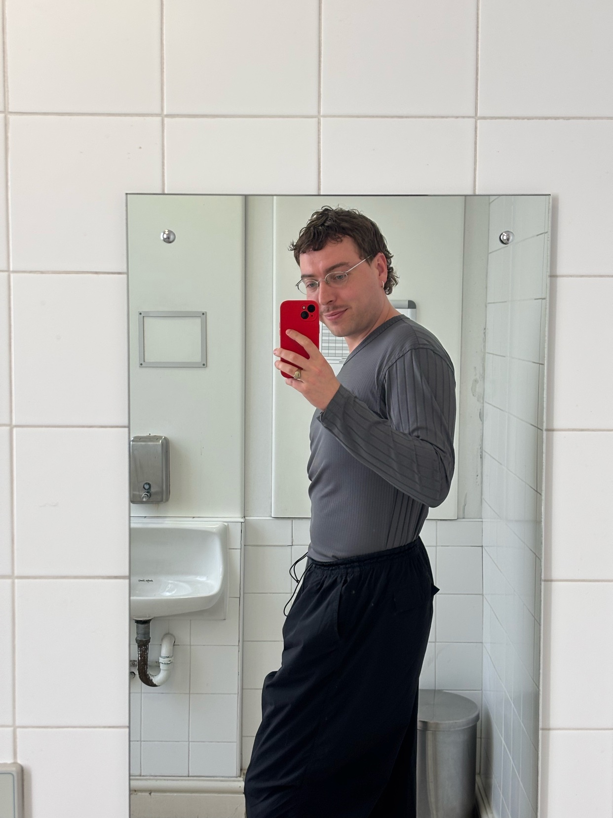 A man in glasses and a grey long-sleeved shirt takes a mirror selfie in a tiled bathroom.