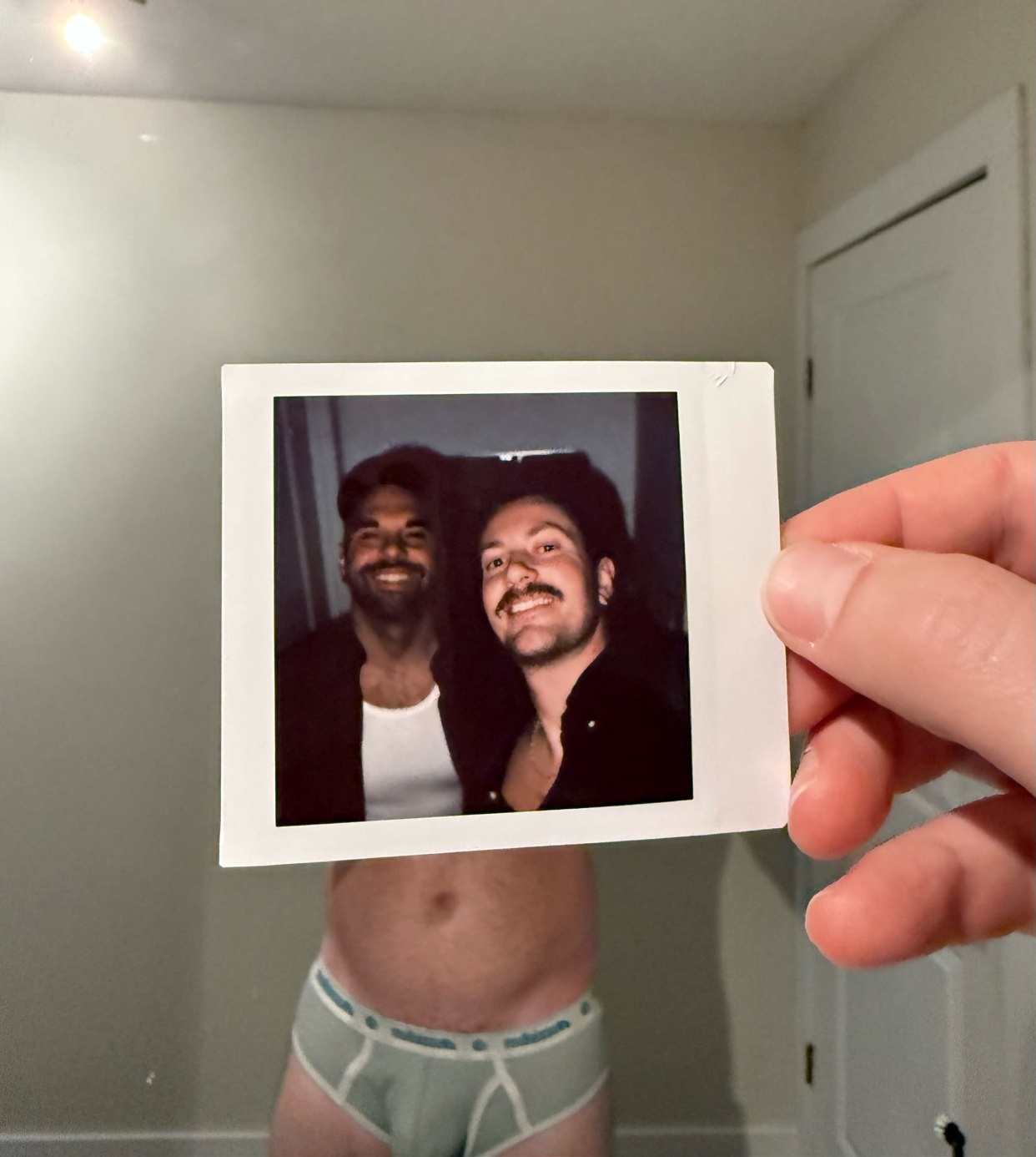 A man holds up a polaroid photo of himself and another man. The man holding the photo is shirtless and wearing underwear.