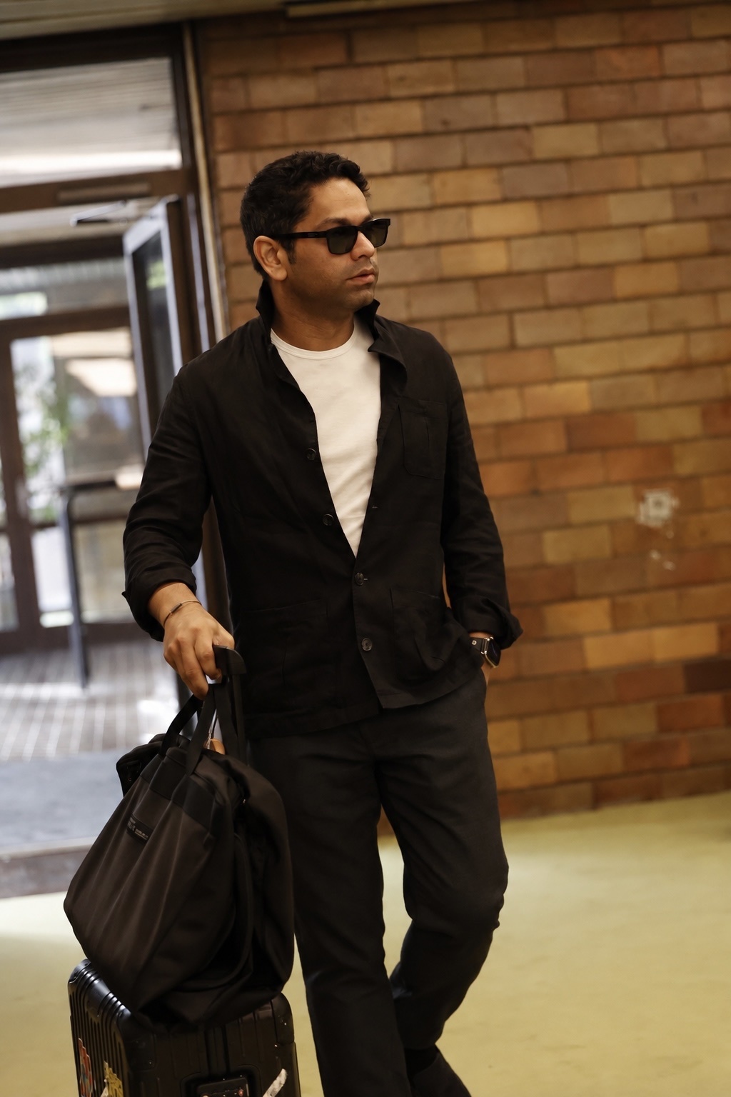 A man in sunglasses and a black button-down shirt over a white t-shirt, holding luggage, appears to be at an airport or travel hub.