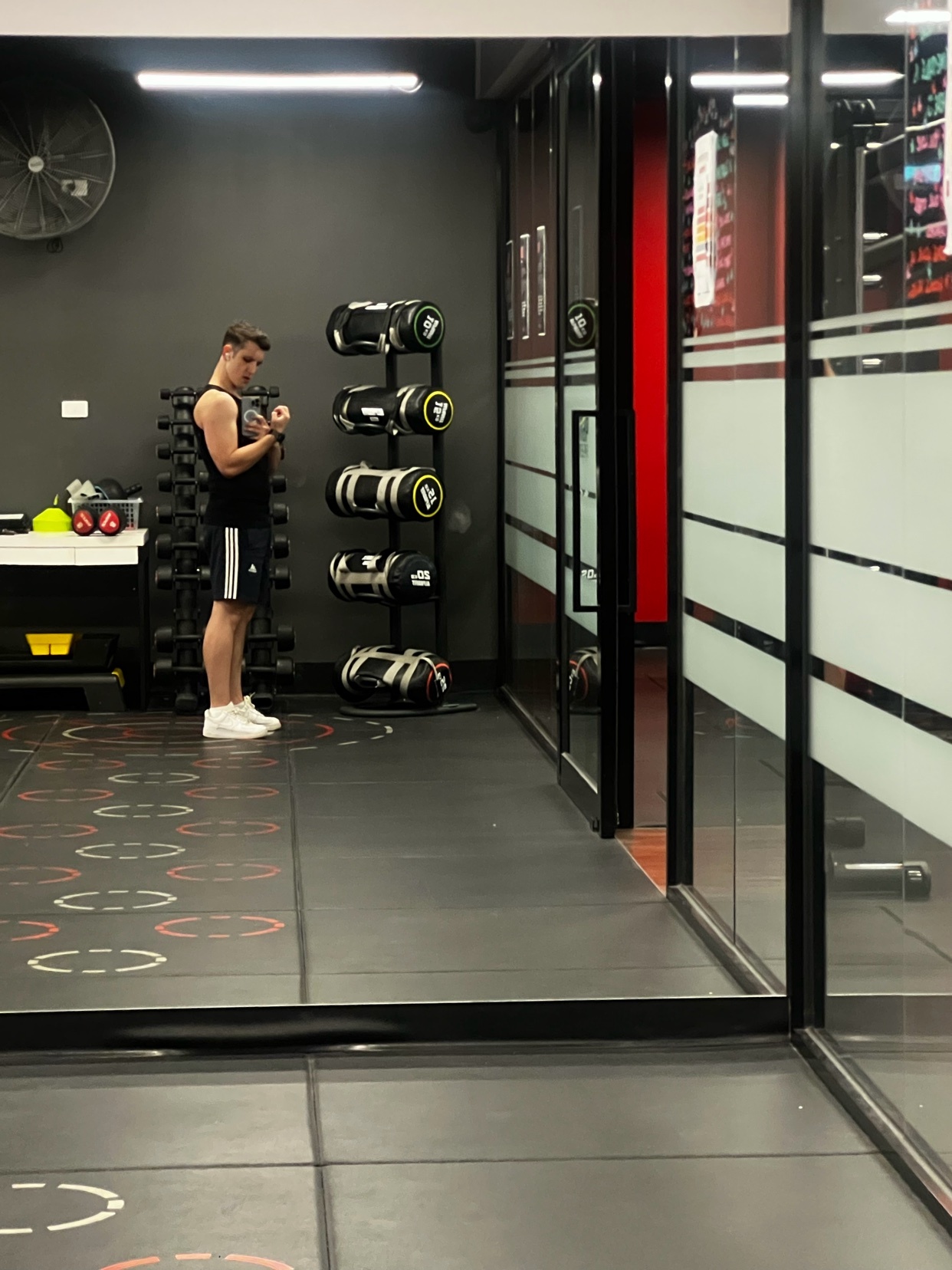 A man stands in a modern gym, looking at his watch. He is wearing athletic shorts and a tank top, with sandbags and other workout equipment visible in the background.