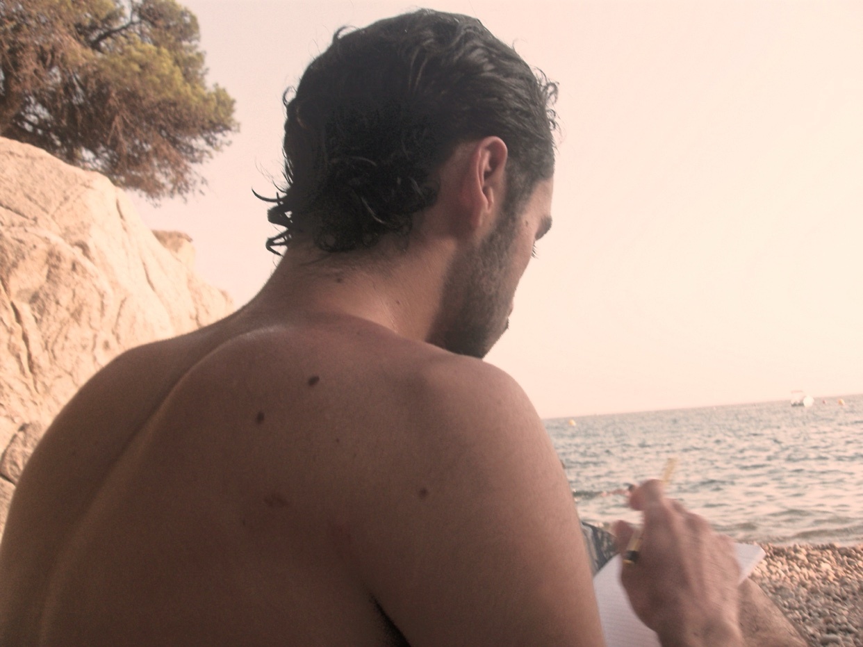 A man sits shirtless on a beach, writing in a notebook. The photo is taken from behind, showing his back and the ocean in the background.