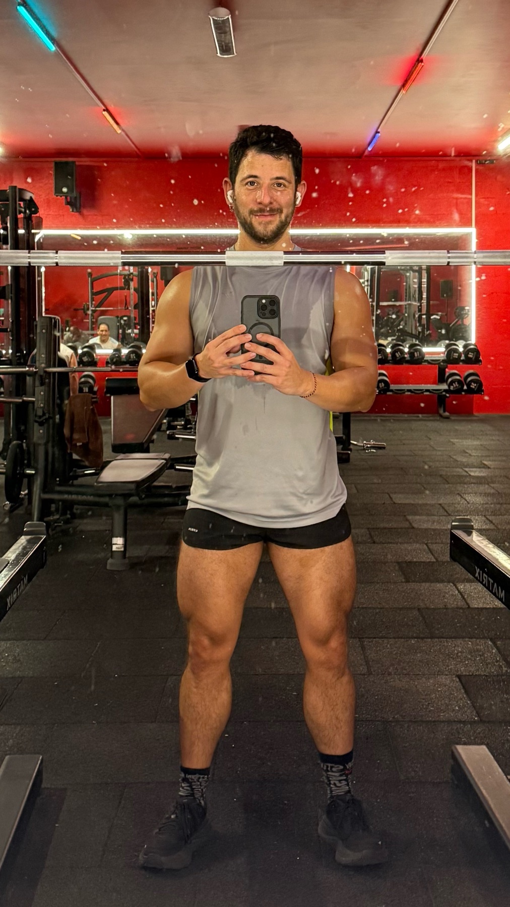 A man takes a mirror selfie in a gym, showcasing his physique. He's wearing a tank top and shorts, highlighting his muscular legs.