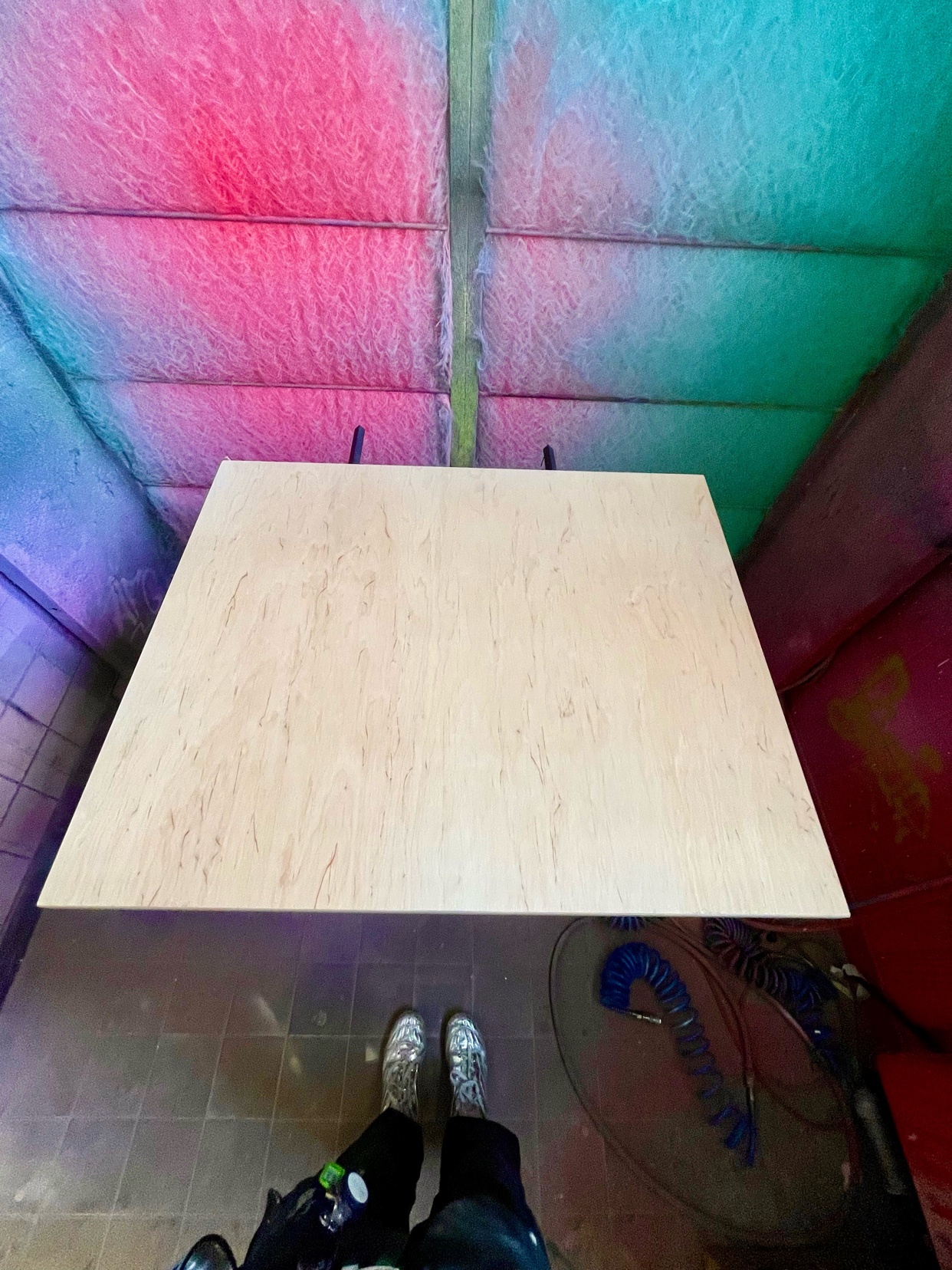 A top-down view of a large piece of plywood resting on a table inside a spray paint booth. The ceiling of the booth is illuminated with pink and teal lights, and the walls are covered in graffiti.