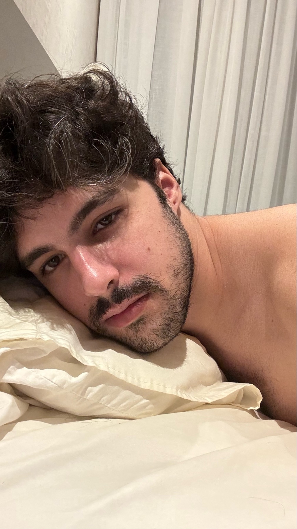 A close-up shot of a man with dark, messy hair and stubble, resting his head on a pillow in bed.