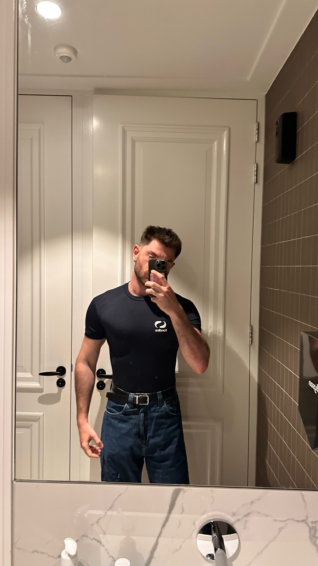 A man takes a mirror selfie in a bathroom, wearing a dark t-shirt and jeans.