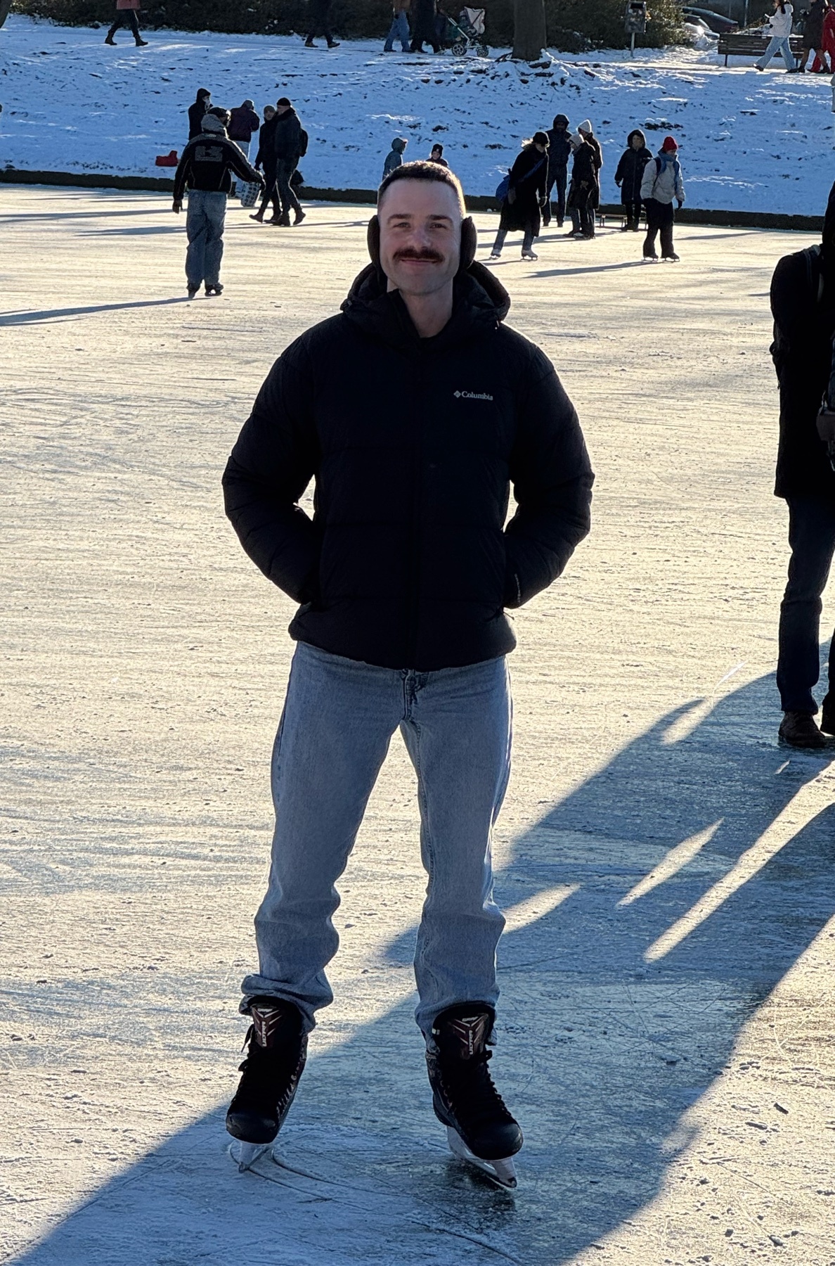 A man with a mustache is ice skating on a frozen lake in a park. He is wearing a black hoodie and jeans, with other people visible in the background enjoying the winter weather.