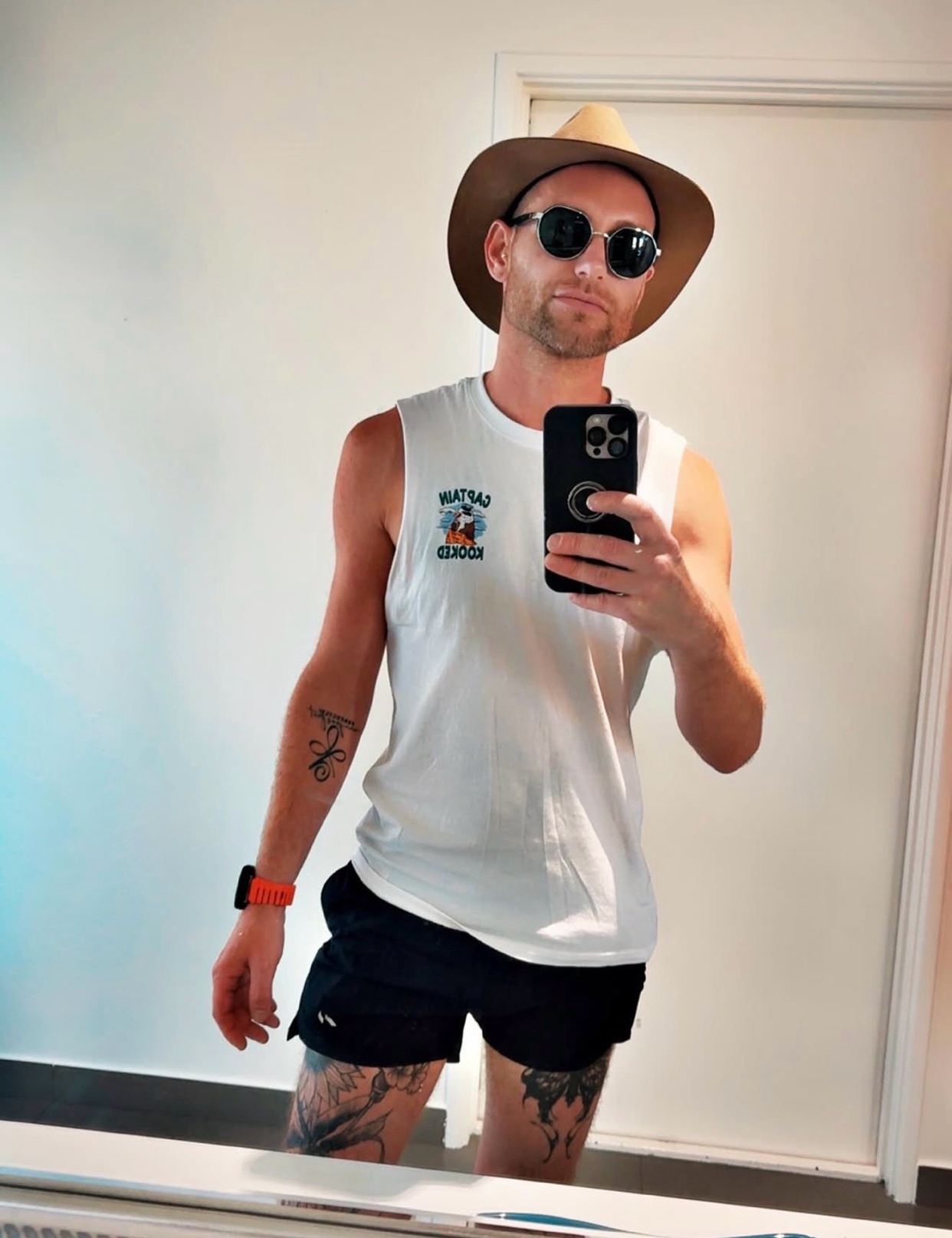 A man in a cowboy hat and sunglasses takes a mirror selfie while wearing a white tank top and black shorts. He has tattoos on his arms and legs.