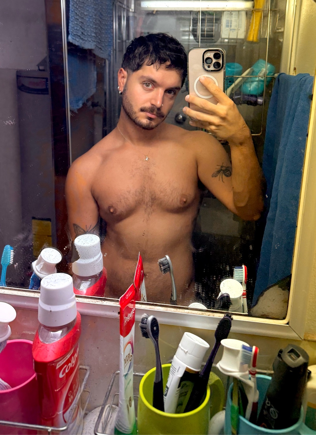 A man takes a mirror selfie in a cluttered bathroom, showing off his tattoos and stubble.