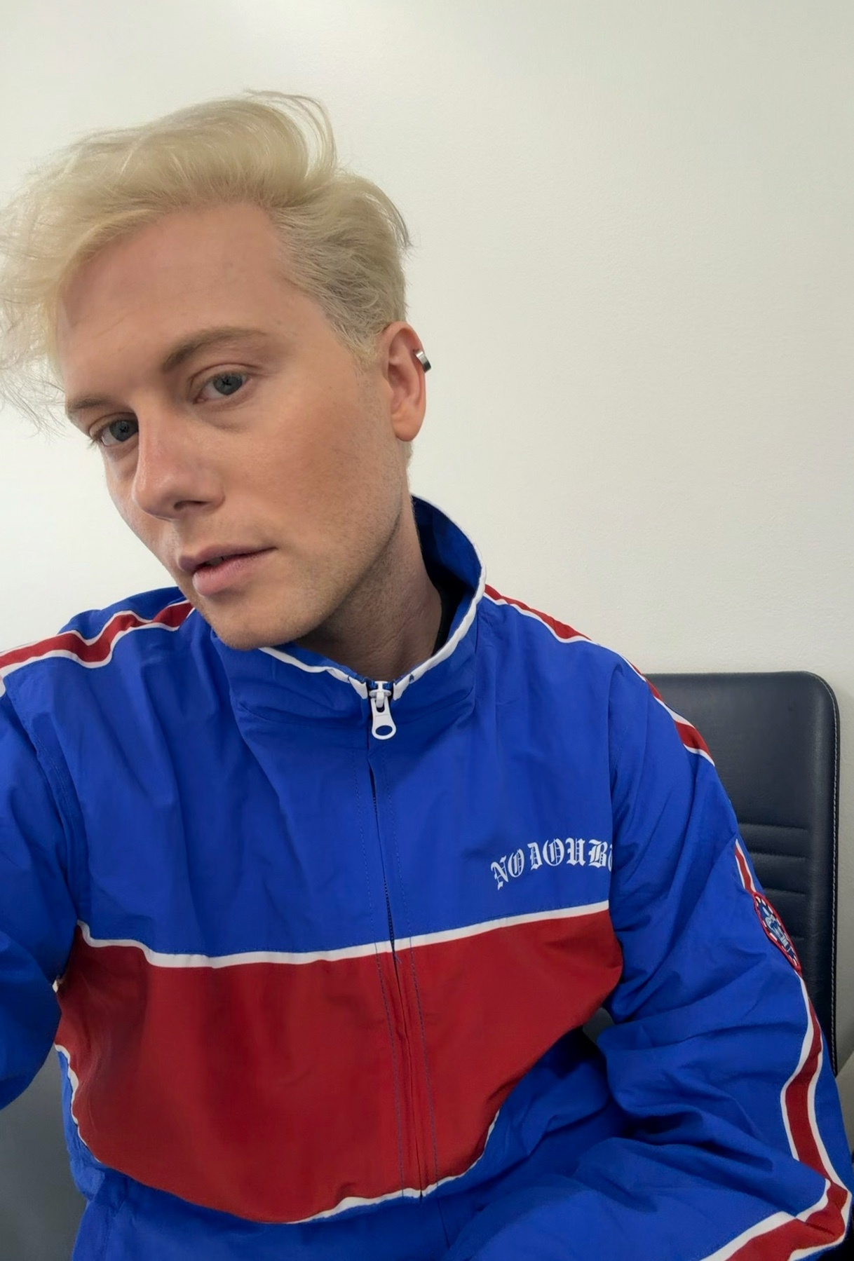 A man with blond hair and blue eyes poses for a selfie wearing a blue, red, and white tracksuit.