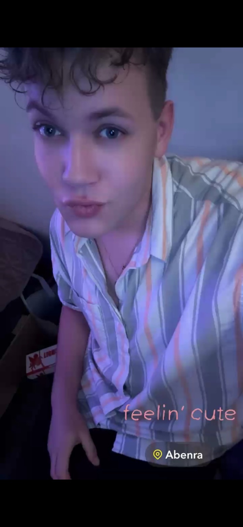 A young man with curly hair and blue eyes poses for a selfie, puckering his lips and wearing a striped button-down shirt. Text overlay reads 'feelin' cute'.