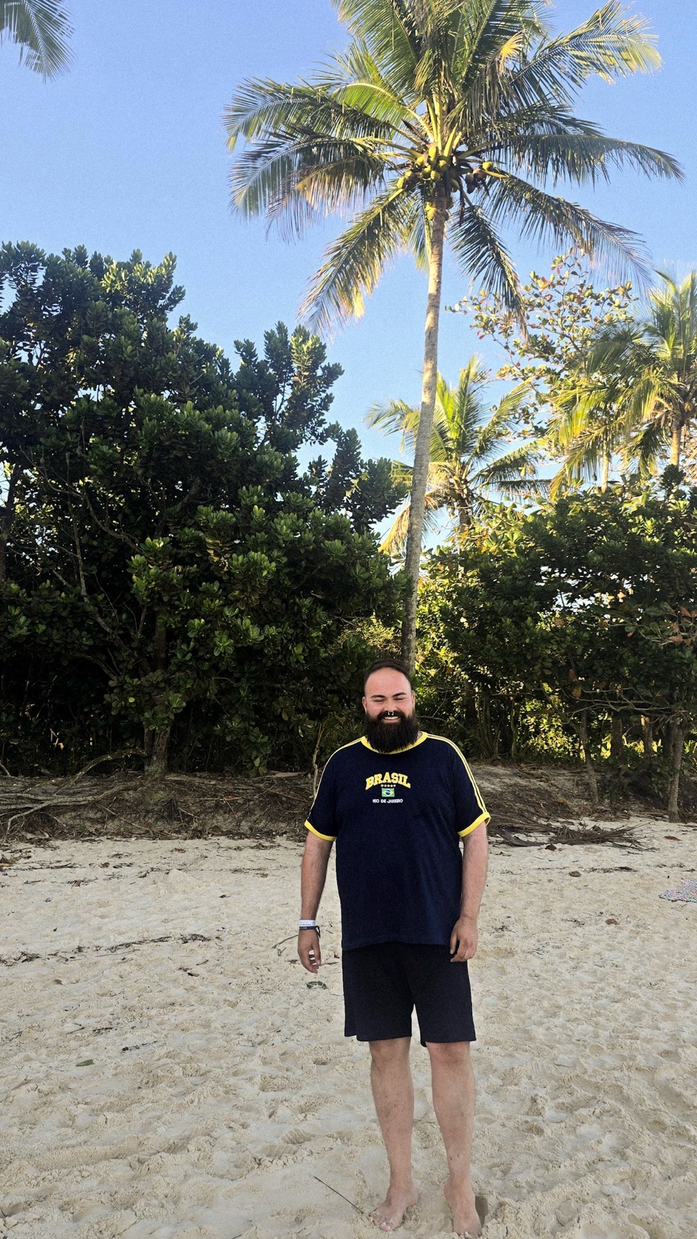A man with a beard stands on a sandy beach with palm trees in the background. The man is wearing a dark blue t-shirt with yellow trim and dark shorts.
