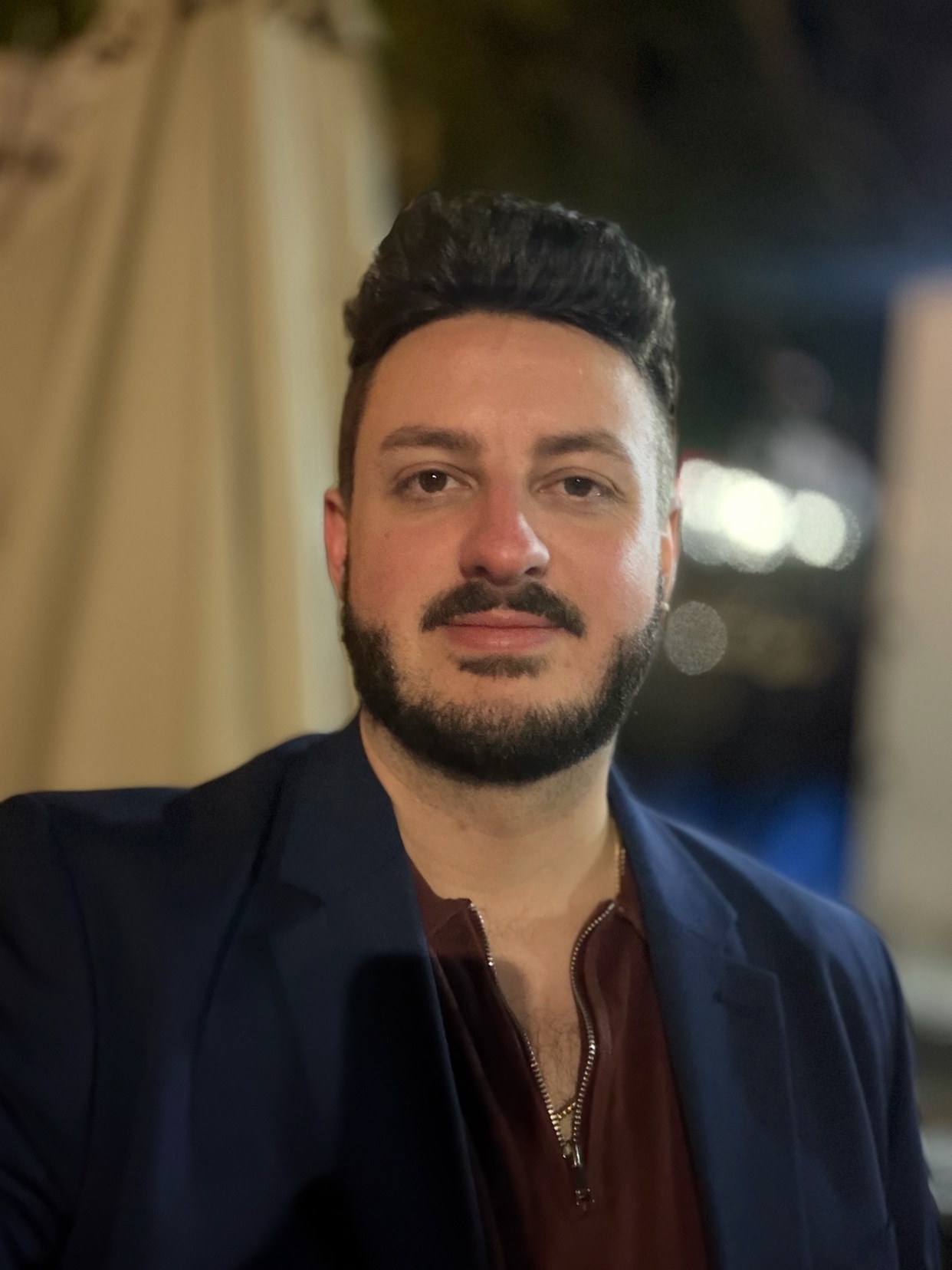 A man with a beard and dark hair poses for a portrait wearing a suit jacket over a maroon shirt.
