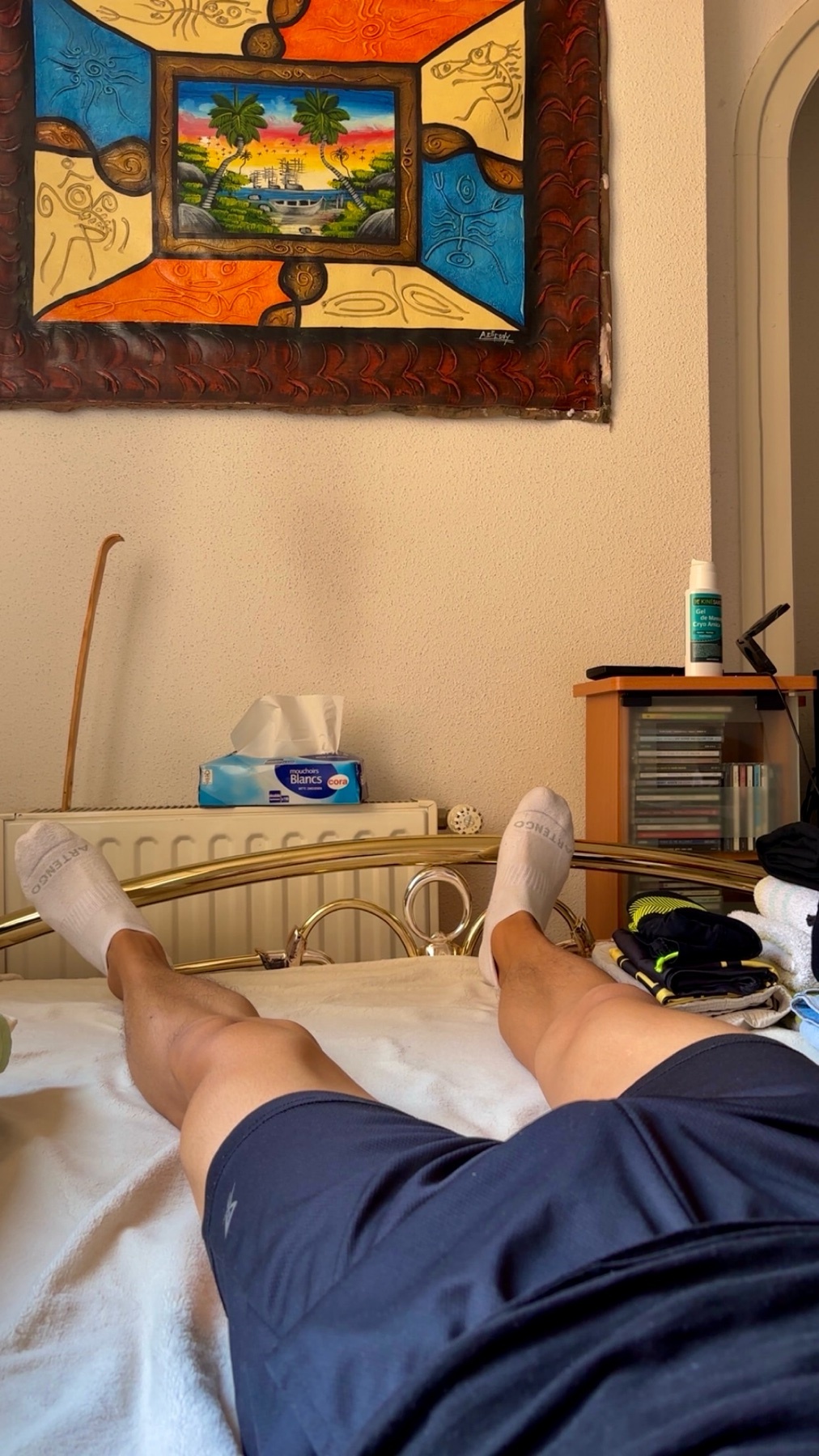 A person's legs are shown from the knees down as they lie on a bed. The person is wearing dark blue shorts and white socks. A colorful framed artwork hangs on the wall above the bed.
