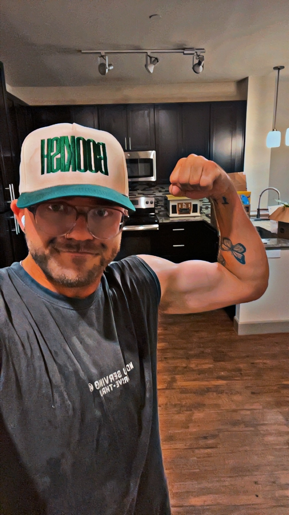 A man with a tattoo on his arm flexes his bicep while wearing a hat and glasses. He is in what appears to be a kitchen or living area.